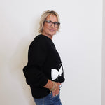 Person wearing a black sweater with a white design and blue jeans against a plain background