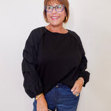 Woman wearing a black top and blue jeans, holding a clutch against a plain background