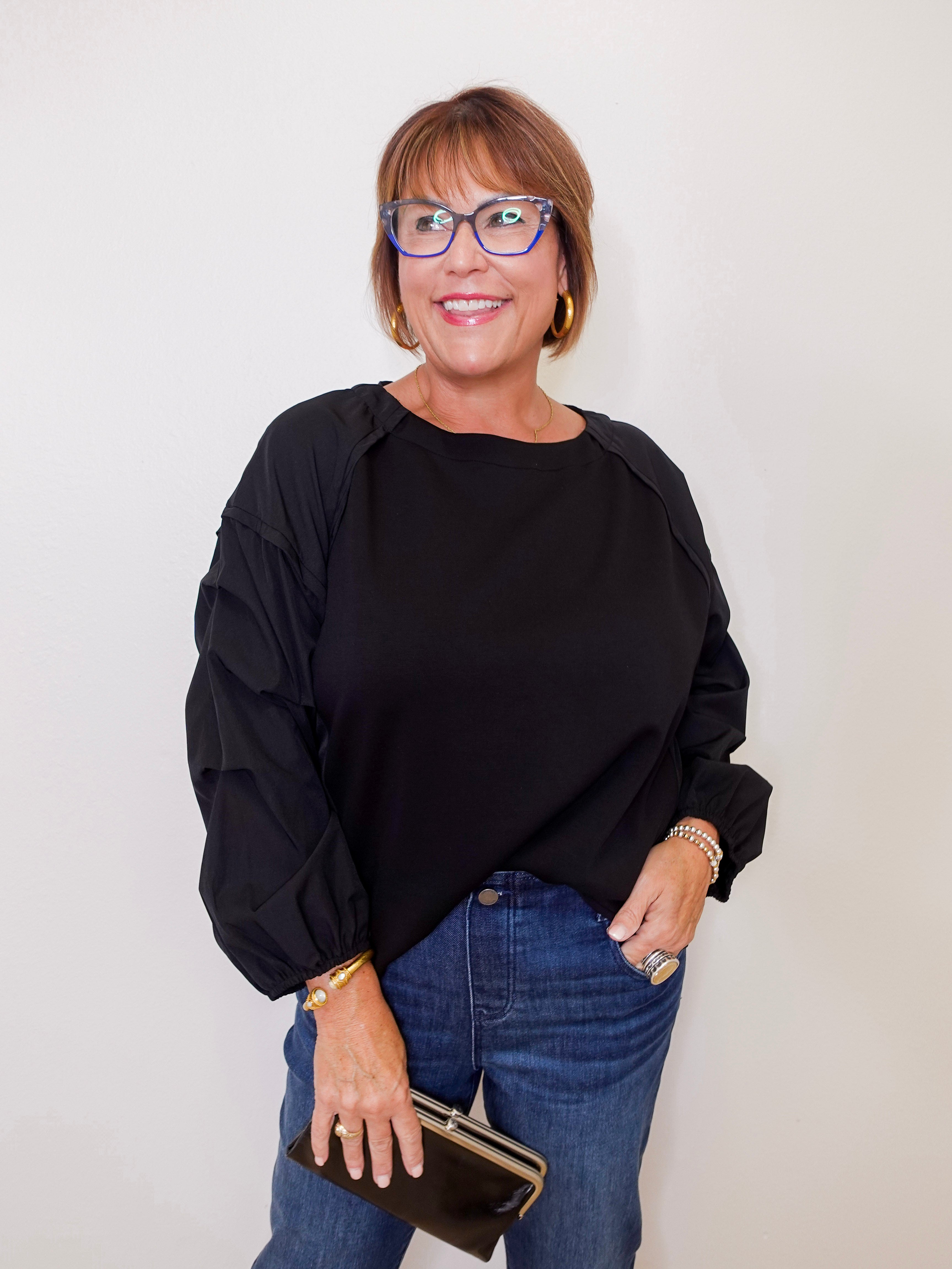 Woman wearing a black top and blue jeans, holding a clutch against a plain background
