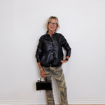 Person wearing a black leather jacket, camouflage pants, and white sneakers against a white wall.