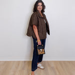 Woman wearing a brown jacket, blue jeans, and white sneakers holding a leopard print handbag against a white wall.