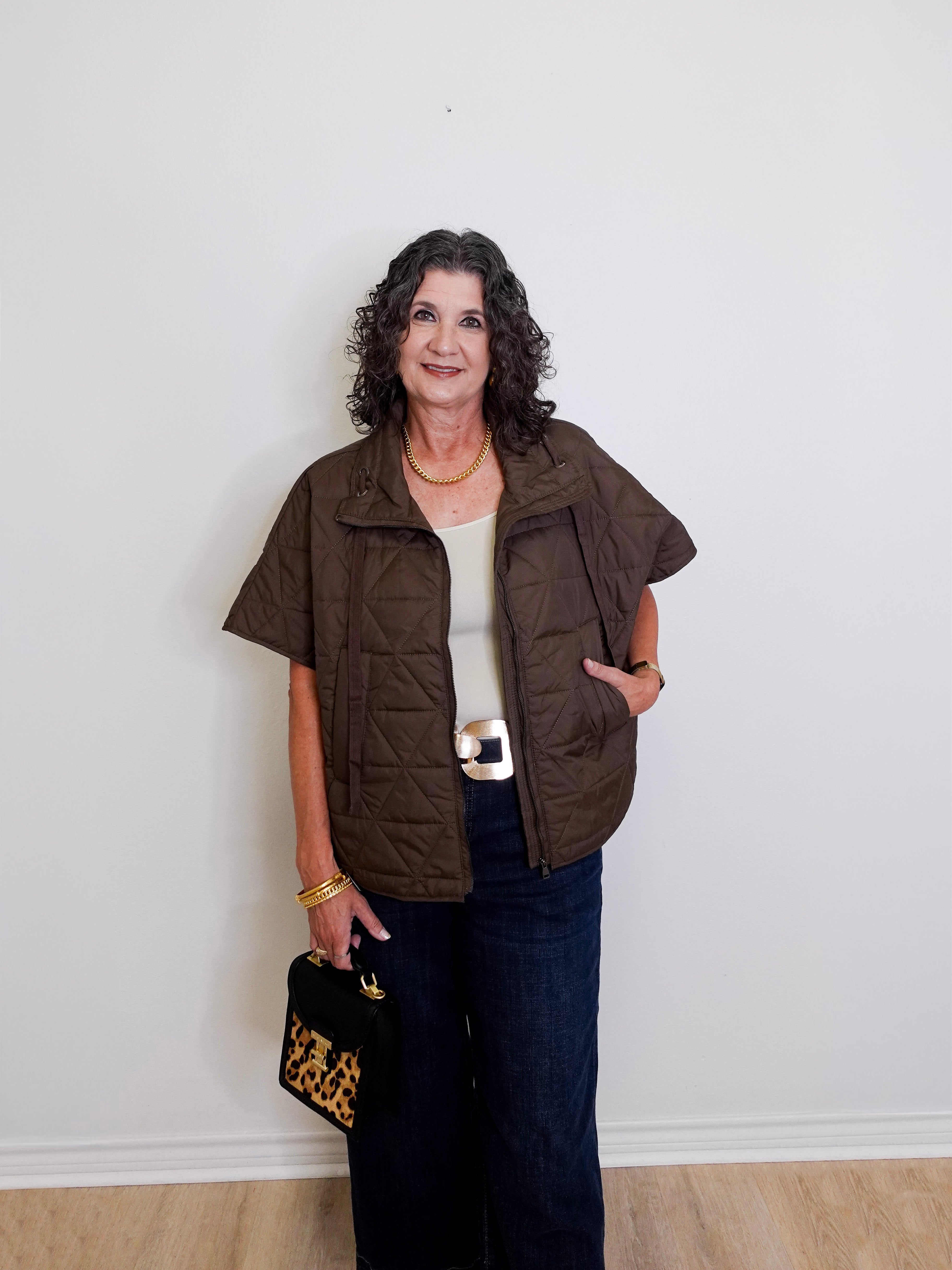 Woman wearing a brown vest over a white top and dark pants, holding a leopard print handbag against a white wall.