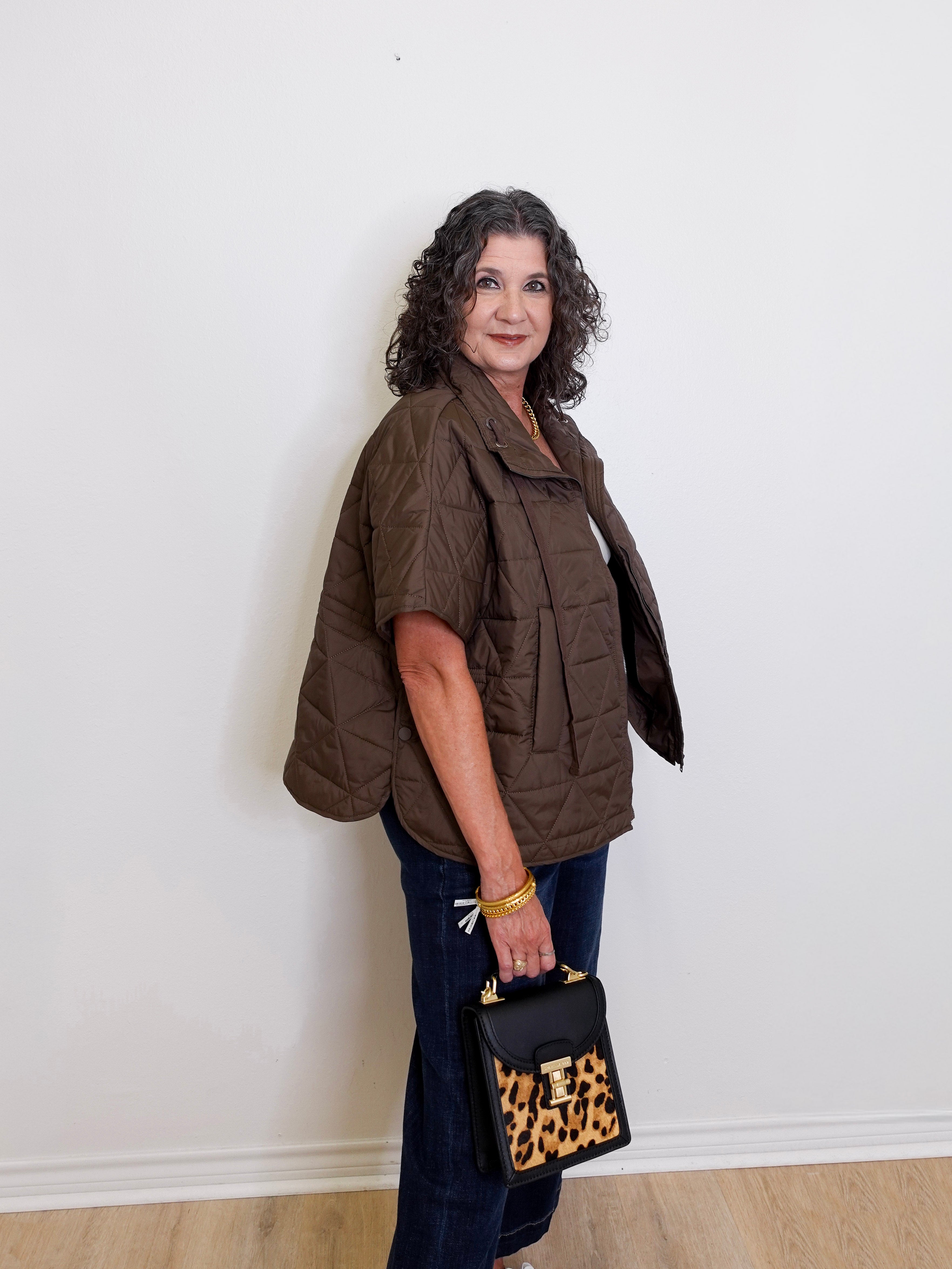 Woman wearing a brown puffer vest and holding a black handbag with leopard print accents against a white wall.
