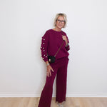 Woman wearing a maroon outfit with pearl details against a white wall.