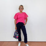 Person wearing a pink top and dark pants, holding a colorful bag against a white wall.