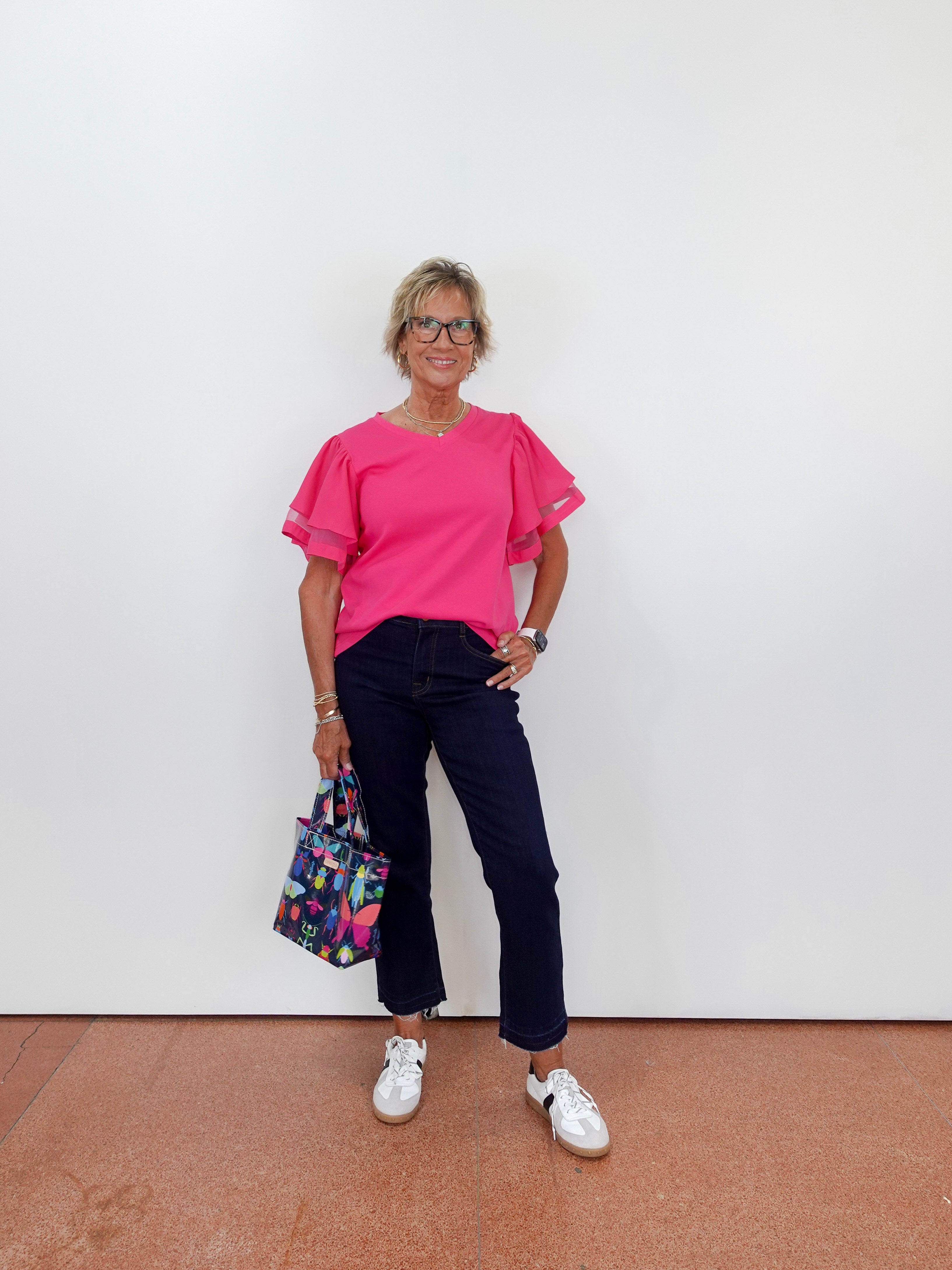 Person wearing a pink top and dark pants, holding a colorful bag against a white wall.