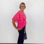 Woman wearing a pink blouse and dark pants against a white wall.