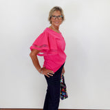 Woman wearing a pink blouse and dark pants against a white wall.