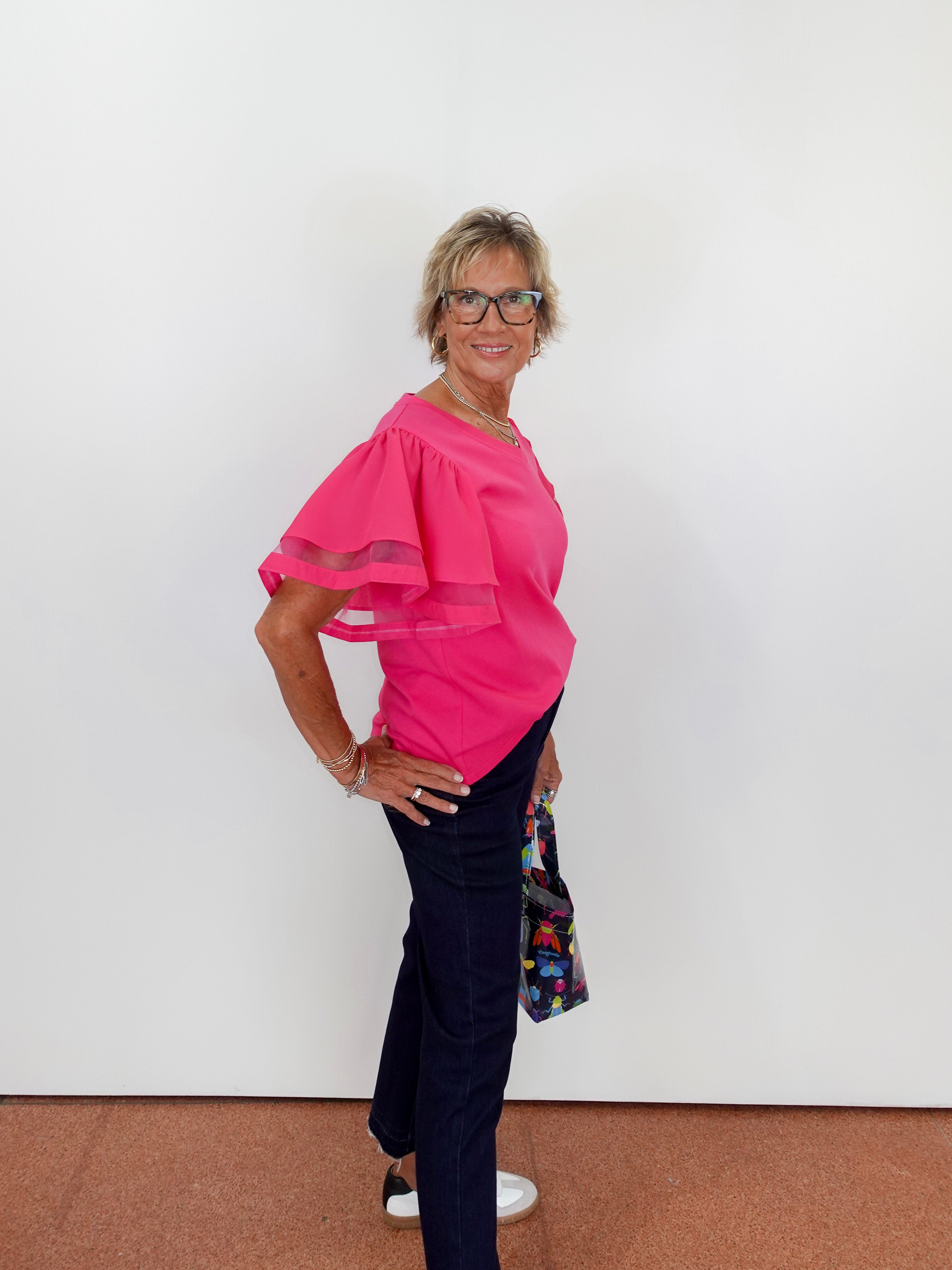 Woman wearing a pink blouse and dark pants against a white wall.
