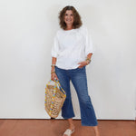 Woman in white blouse and blue jeans holding a patterned bag against a white wall.