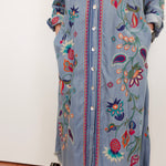 A light blue denim shirt dress with colorful floral embroidery and button-front closure, shown on a person standing against a plain indoor wall.