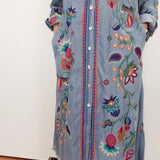 A light blue denim shirt dress with colorful floral embroidery and button-front closure, shown on a person standing against a plain indoor wall.