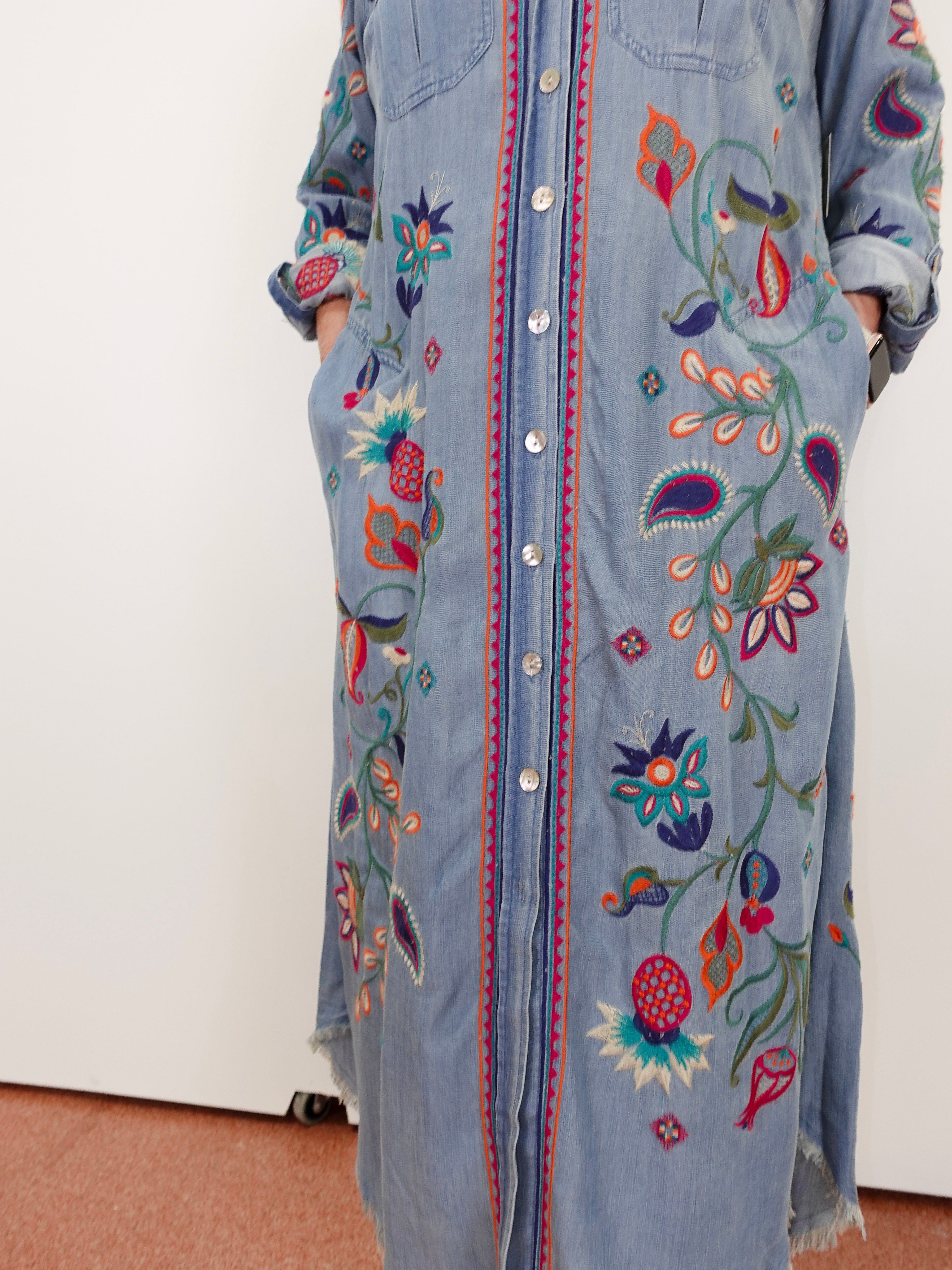 A light blue denim shirt dress with colorful floral embroidery and button-front closure, shown on a person standing against a plain indoor wall.