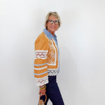 Woman wearing a mustard yellow jacket with lace details against a white wall.
