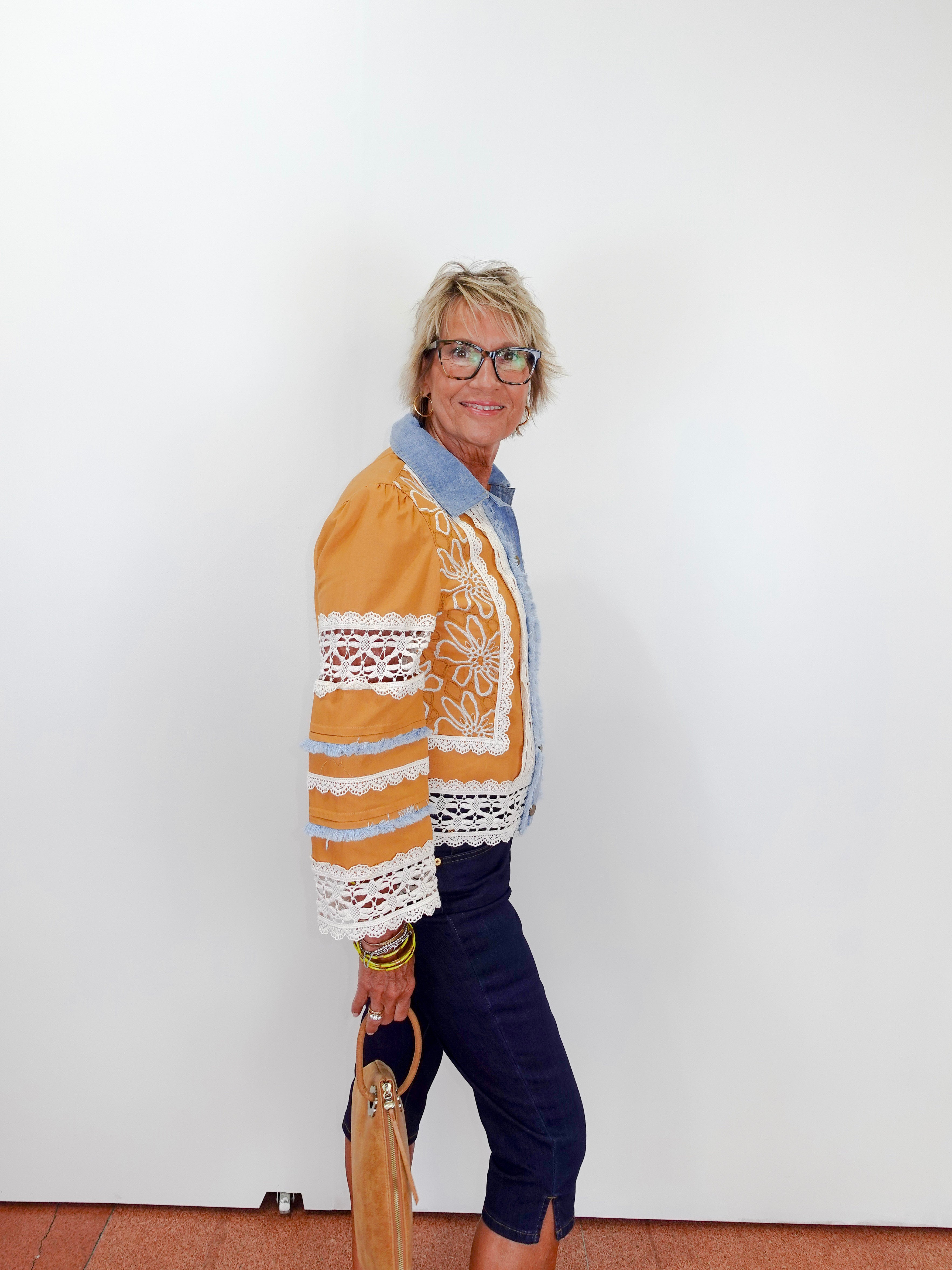 Woman wearing a mustard yellow jacket with lace details against a white wall.