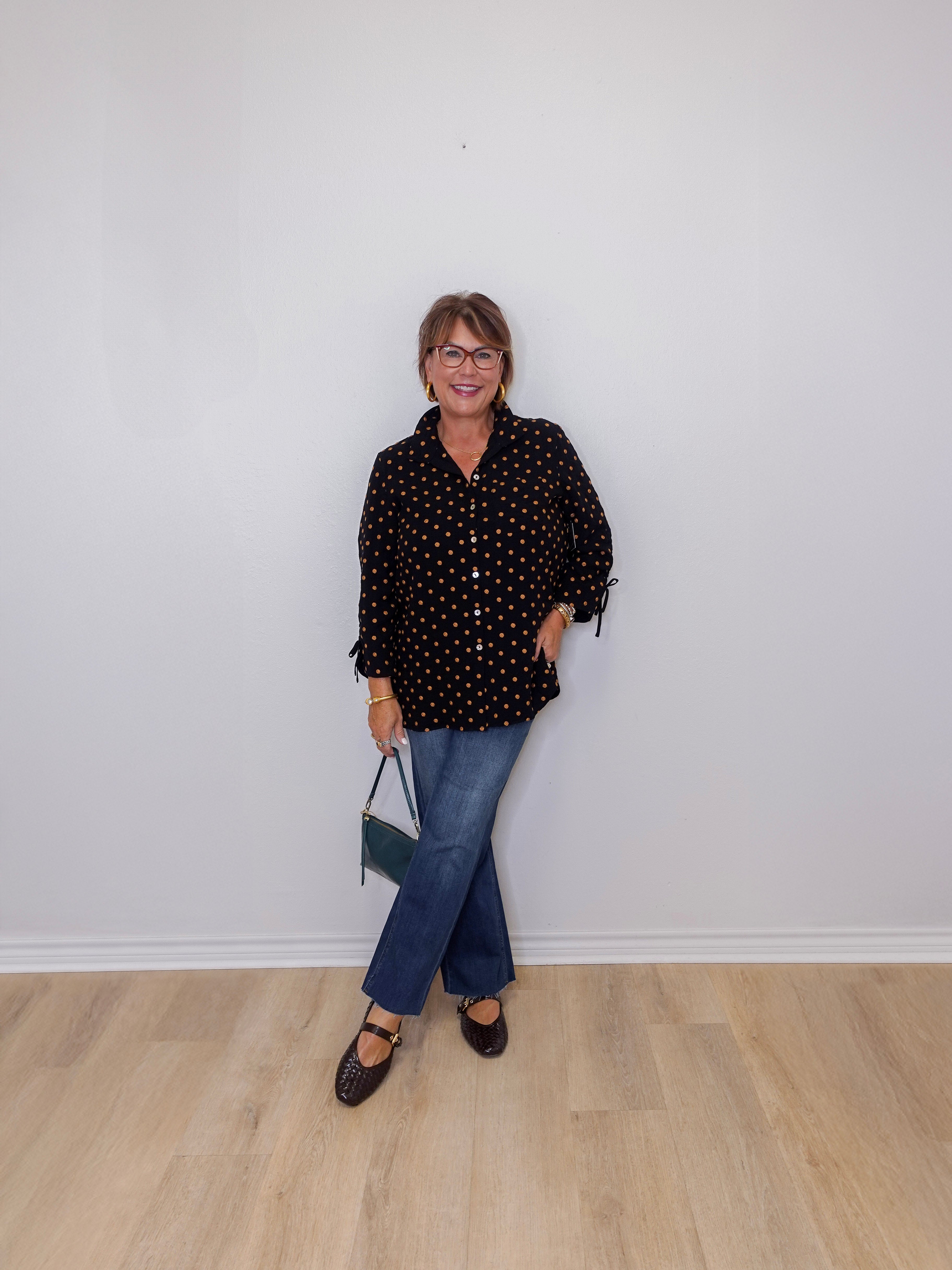 Person wearing a black polka dot shirt and blue jeans against a white wall.