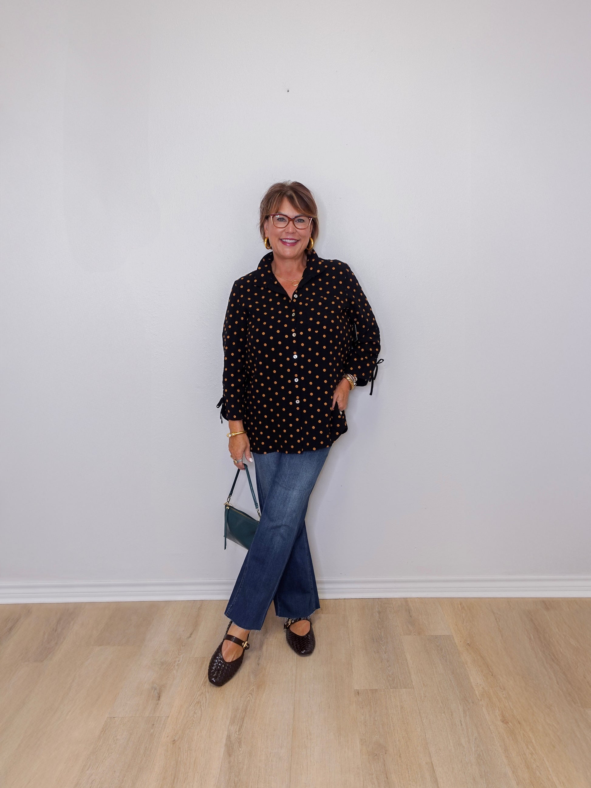 Person wearing a black polka dot shirt and blue jeans against a white wall.