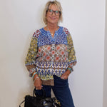 Woman wearing a patterned blouse and holding a black handbag against a white wall.