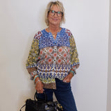 Woman wearing a patterned blouse and holding a black handbag against a white wall.