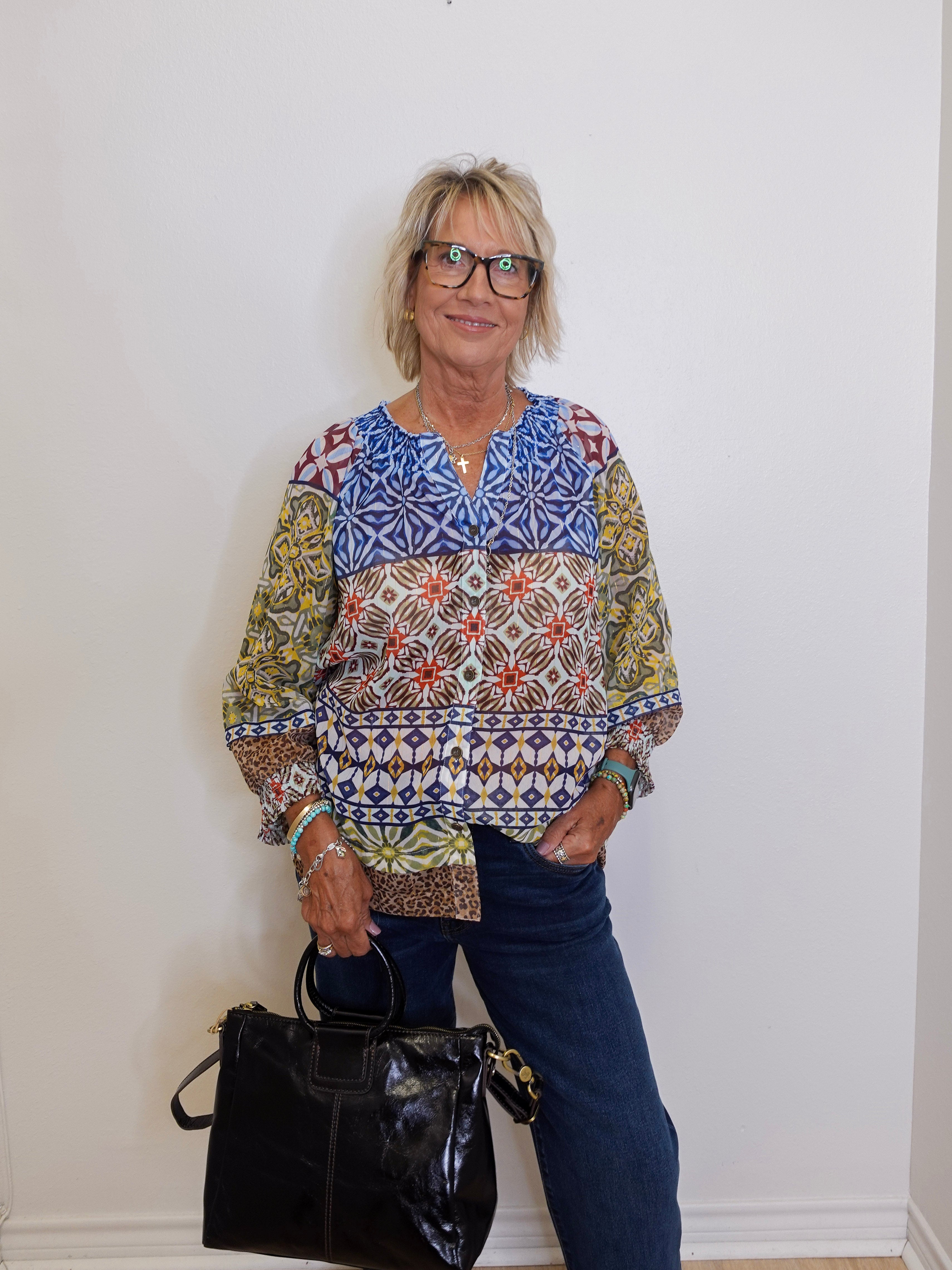 Woman wearing a patterned blouse and holding a black handbag against a white wall.