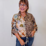 Woman wearing a leopard print shirt over a floral blouse with blue jeans against a white wall.