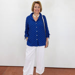 Woman wearing a blue polka dot blouse and white pants against a white wall.