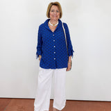 Woman wearing a blue polka dot blouse and white pants against a white wall.