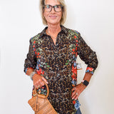 Woman wearing a leopard print shirt with floral sleeves, holding a brown handbag against a white background.