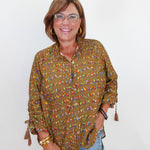 A woman wearing a long-sleeve button-up blouse with a busy multicolor bird print on an olive green base, paired with light blue jeans and gold jewelry, standing against a plain white background.