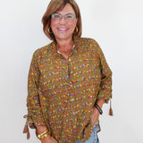 A woman wearing a long-sleeve button-up blouse with a busy multicolor bird print on an olive green base, paired with light blue jeans and gold jewelry, standing against a plain white background.