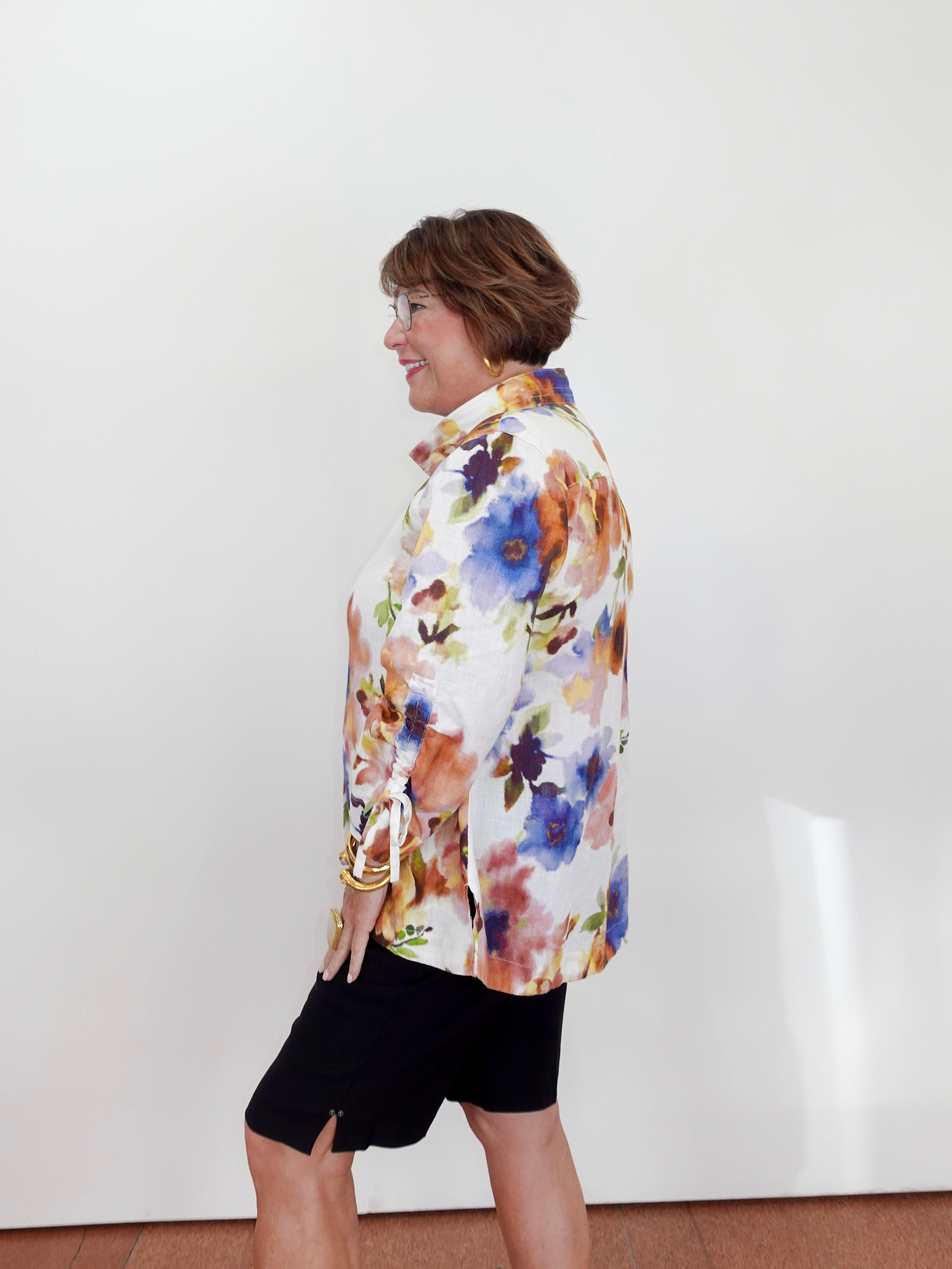 Woman wearing a colorful floral blouse and black shorts against a white wall.