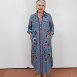 An older woman stands against a plain white wall wearing a long blue denim shirt dress with colorful floral embroidery and button-front closure, paired with tan slide sandals.
