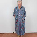 An older woman stands against a plain white wall wearing a long blue denim shirt dress with colorful floral embroidery and button-front closure, paired with tan slide sandals.
