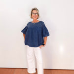 Woman wearing a blue blouse and white pants standing against a white wall.