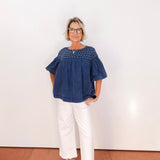 Woman wearing a blue blouse and white pants standing against a white wall.