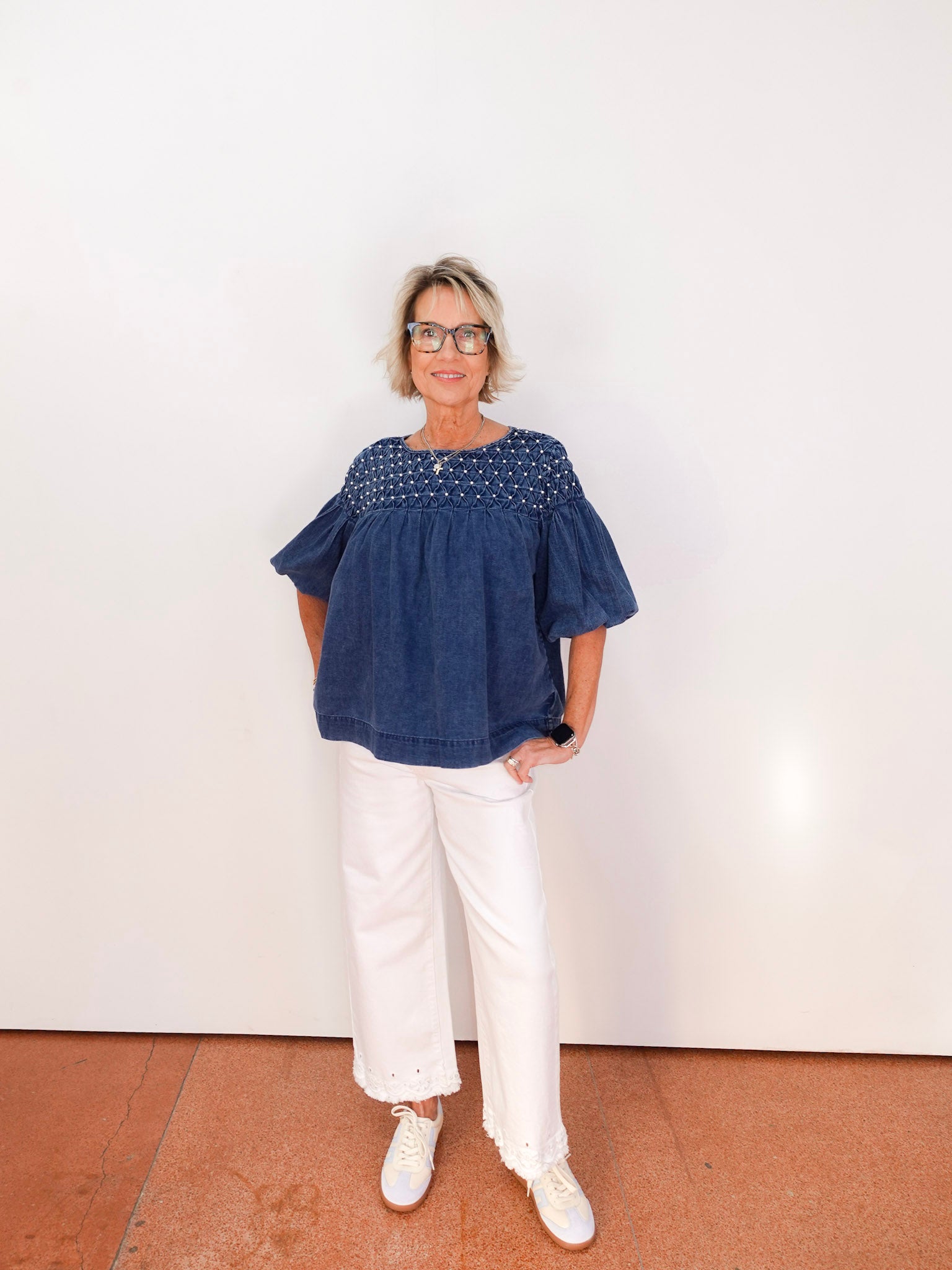 Woman wearing a blue blouse and white pants standing against a white wall.