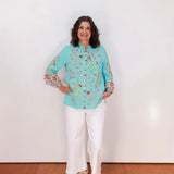 Woman wearing a light blue floral blouse and white pants against a white wall.
