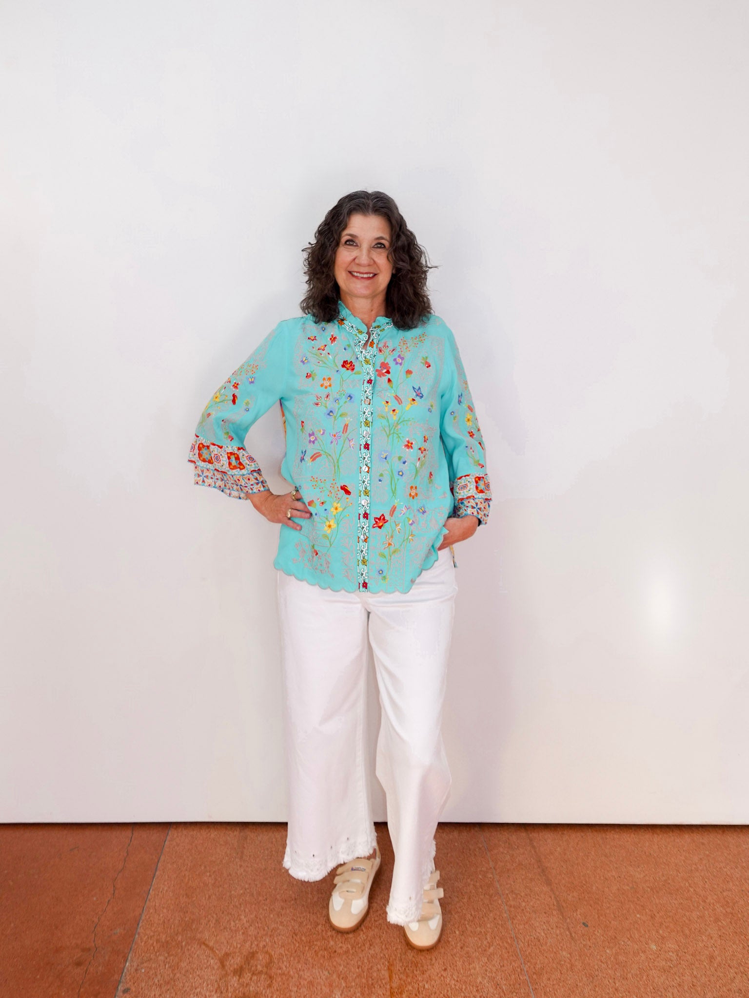 Woman wearing a light blue floral blouse and white pants against a white wall.