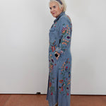 A woman stands in profile wearing a long light blue denim-style coat with colorful floral and bird embroidery, paired with tan sandals and a dark headband, against a plain white wall.