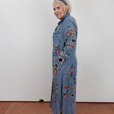 A woman stands in profile wearing a long light blue denim-style coat with colorful floral and bird embroidery, paired with tan sandals and a dark headband, against a plain white wall.