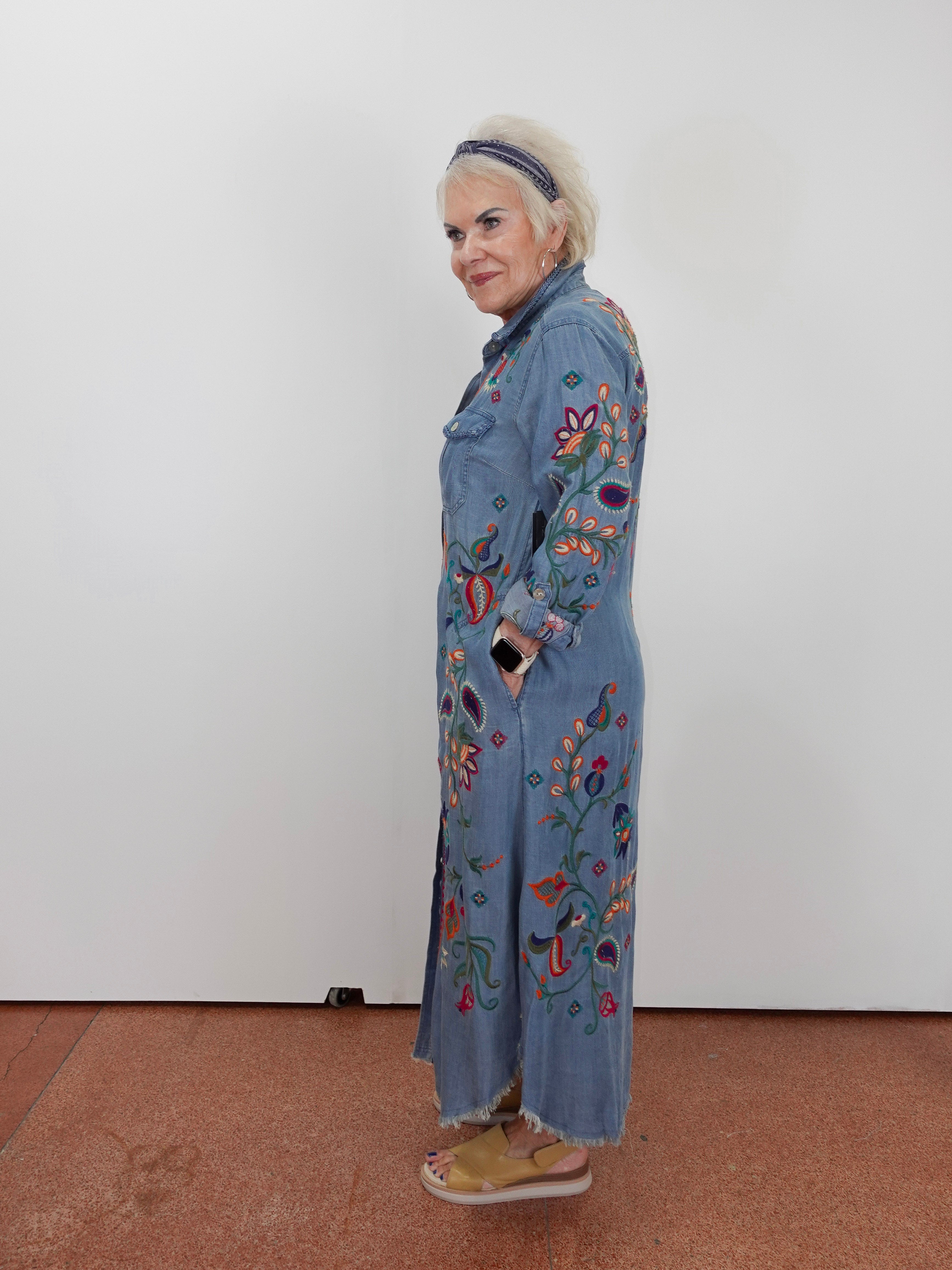 A woman stands in profile wearing a long light blue denim-style coat with colorful floral and bird embroidery, paired with tan sandals and a dark headband, against a plain white wall.