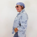 Woman wearing a light blue denim jacket with dog patterns, sunglasses, and a headscarf against a white background.