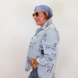 Woman wearing a light blue denim jacket with dog patterns, sunglasses, and a headscarf against a white background.