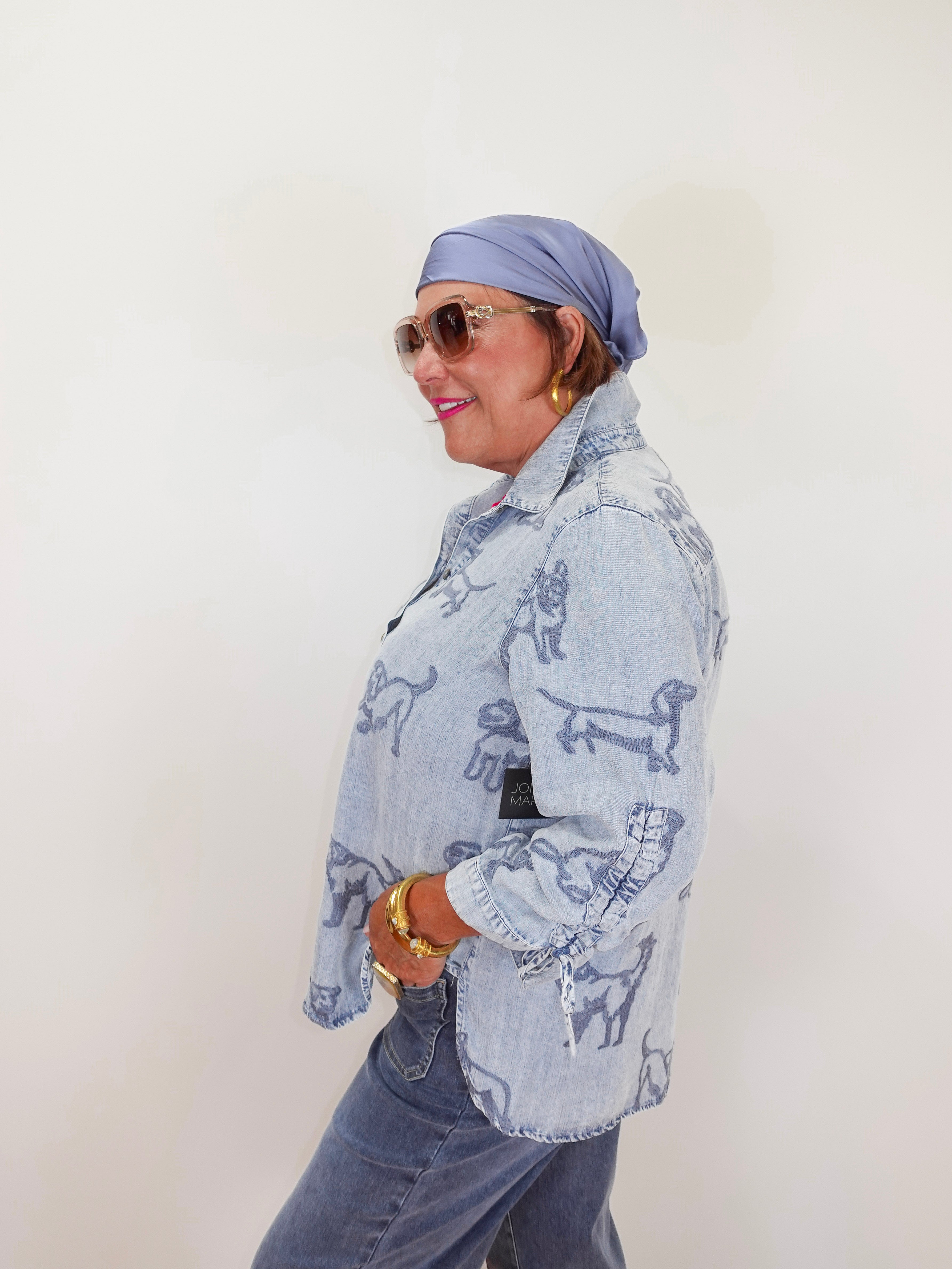 Woman wearing a light blue denim jacket with dog patterns, sunglasses, and a headscarf against a white background.