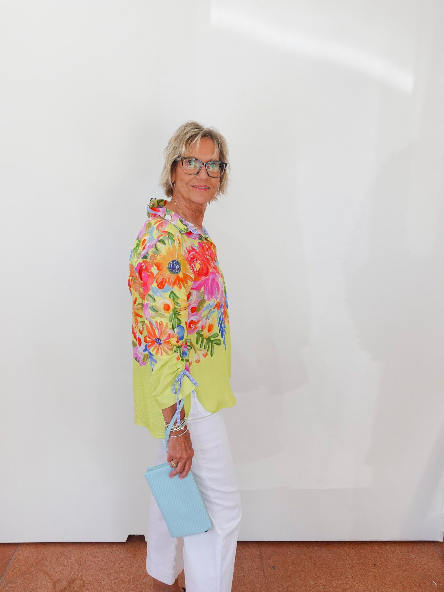 Woman wearing a colorful floral blouse and white pants against a plain background