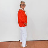 Person wearing an orange top and white pants standing against a white wall.