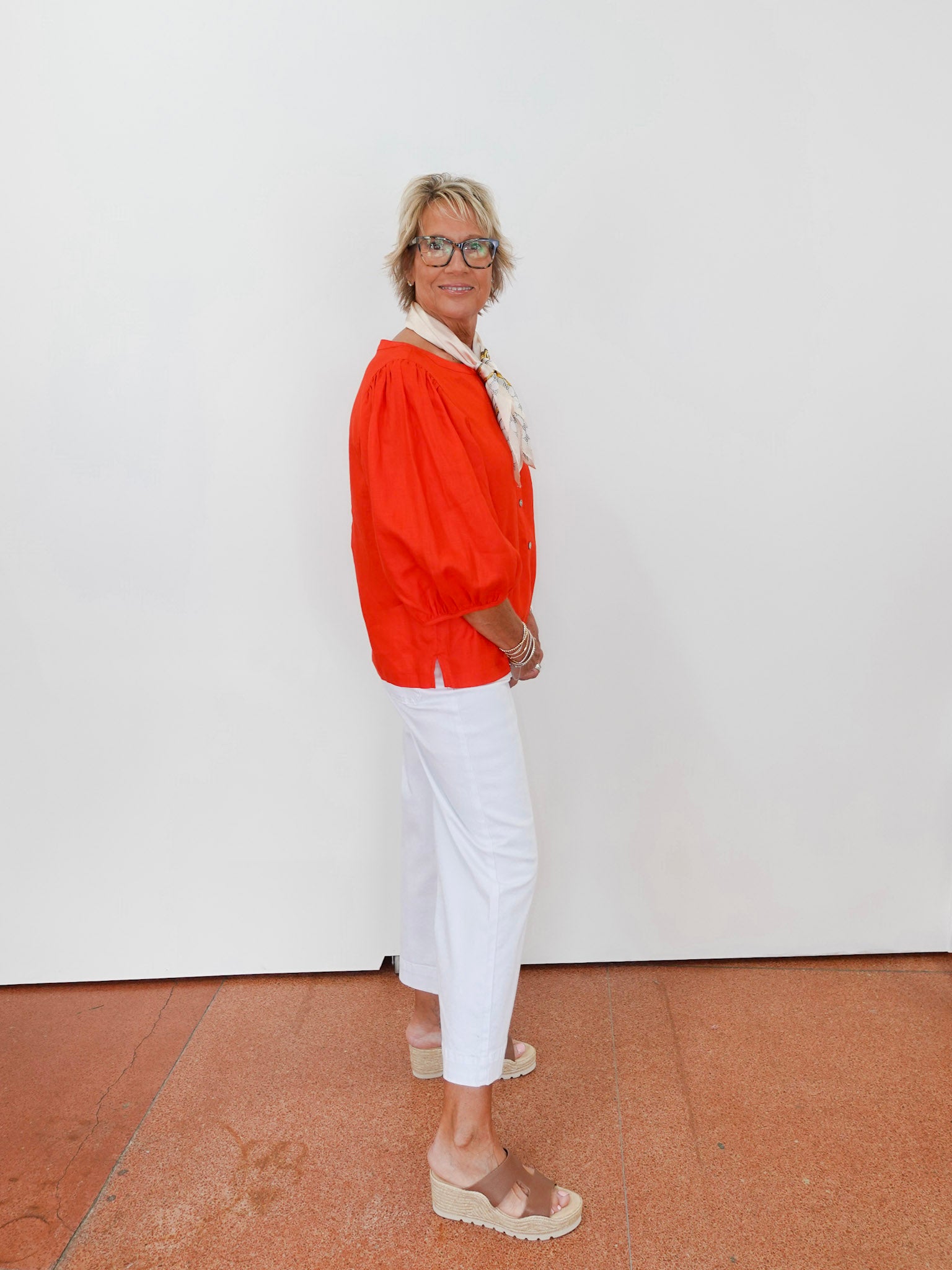 Person wearing an orange top and white pants standing against a white wall.