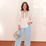 Woman wearing a white blouse with red embroidery and light blue pants, standing against a plain background.