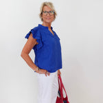 Woman wearing a blue top with ruffled sleeves and white pants, holding a red handbag against a white background.