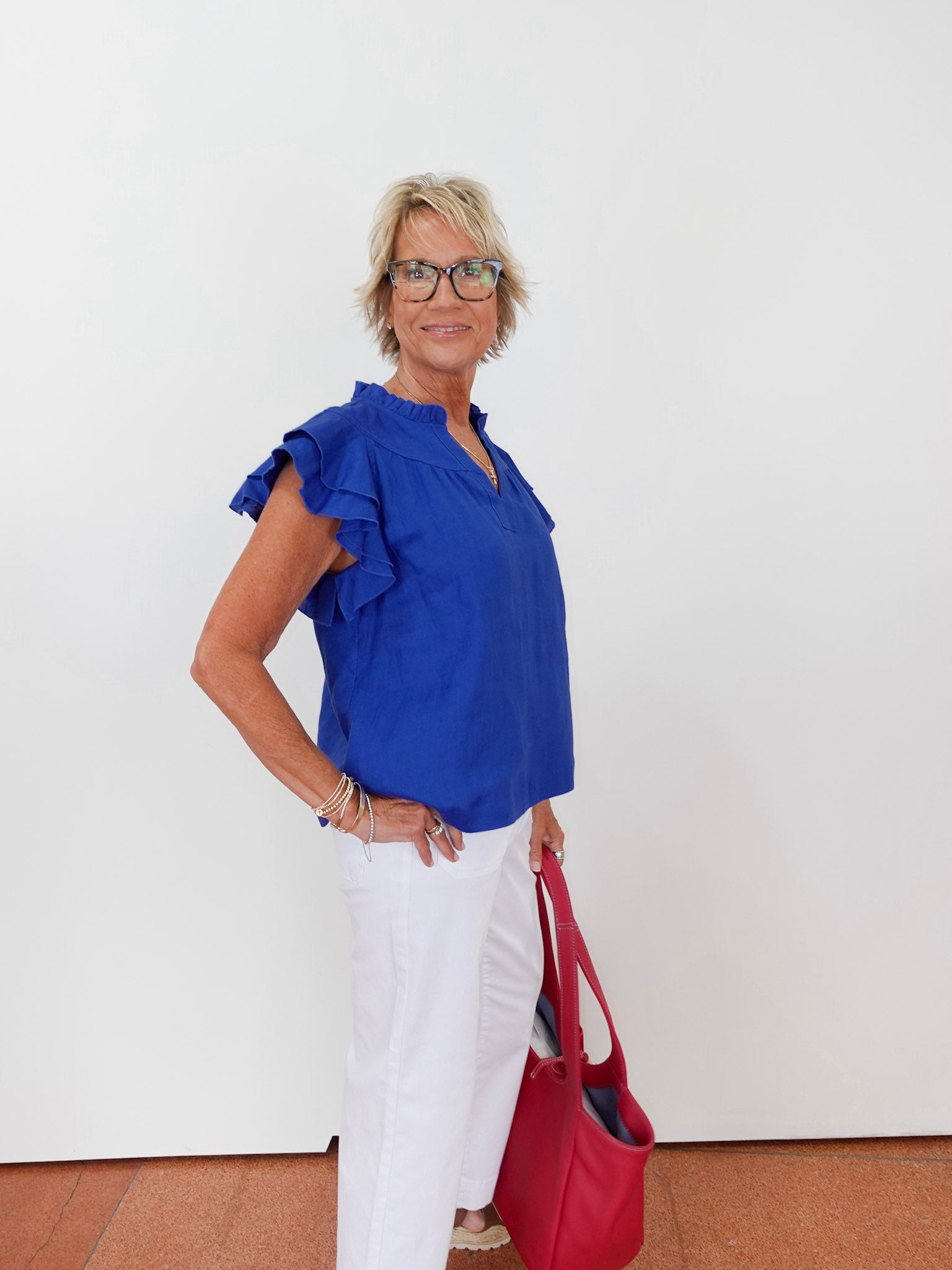 Woman wearing a blue top with ruffled sleeves and white pants, holding a red handbag against a white background.